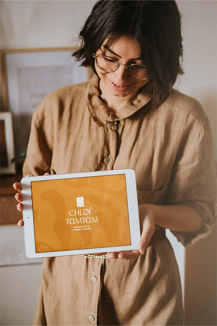 Portrait de Chloé Leonelli, fondatrice et designer de marque et graphiste éco-responsable de Chloé TamTam, tenant dans ses mains une tablette avec pour fond d'écran le logo du studio.